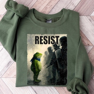 May include: An olive green sweatshirt featuring a graphic with the word "RESIST" above an illustration of a frog facing a line of figures in riot gear. The frog wears a blue scarf. The sweatshirt has long sleeves.