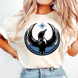 May include: Cream-colored t-shirt featuring a black bird design with outstretched wings, a red eye, and a blue star above. The bird is encircled by a blue ring. The shirt is paired with light blue denim jeans.