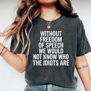 May include: A charcoal grey short-sleeved t-shirt featuring white text that reads: WITHOUT FREEDOM OF SPEECH WE WOULD NOT KNOW WHO THE IDIOTS ARE. The shirt has a crew neckline and a vintage-style washed finish, paired with blue denim jeans.