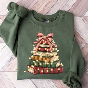 May include: An olive green sweatshirt featuring a stack of books decorated as a Christmas tree. The books are in various colors, with a red and white plaid bow at the top and string lights. The sweatshirt has long sleeves and a crew neck.
