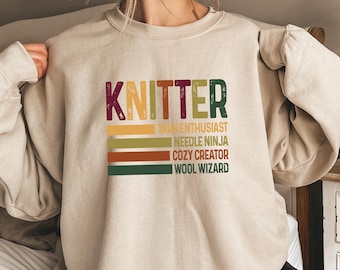 May include: A beige sweatshirt with the word "KNITTER" in colorful letters. Below the word are horizontal stripes in yellow, green, orange, and brown, with the phrases "Yarn Enthusiast", "Needle Ninja", "Cozy Creator", and "Wool Wizard".
