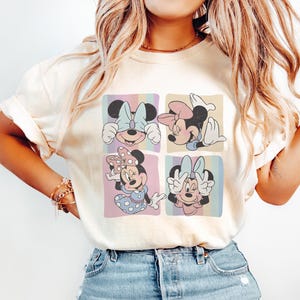 Op de afbeelding: Cr&egrave;mekleurig T-shirt met een vierpaneels afbeelding van Minnie Mouse in verschillende poses. Elk paneel heeft een pastelkleurige achtergrond met een ander Minnie Mouse-ontwerp. Het shirt wordt gecombineerd met lichtblauwe jeans.