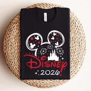 May include: Black t-shirt featuring a white and red Disney design. The design includes Mickey Mouse ears, stars, fireworks, a castle, and the word "Disney" in red, along with the year "2026". The shirt is displayed on a woven surface.
