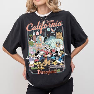 Op de afbeelding: Zwart T-shirt met een kleurrijke afbeelding van Disneyland-personages en -bezienswaardigheden. Het ontwerp bevat de tekst "California Disneyland Est. 1955". Het shirt heeft een relaxte pasvorm en een wit ondershirt komt tevoorschijn.