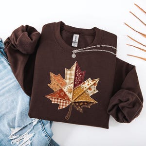 Peut inclure: Un sweat-shirt à col rond marron foncé orné d'une grande applique centrale en forme de feuille d'érable, composée de divers empiècements de tissu aux tons orange, beige et marron avec des motifs floraux et à carreaux. Un collier en argent.