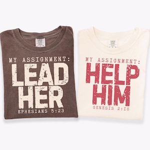 May include: Two folded t-shirts. One is brown with the text "MY ASSIGNMENT: LEAD HER" and "EPHESIANS 5:23". The other is cream-colored with the text "MY ASSIGNMENT: HELP HIM" and "GENESIS 2:18".