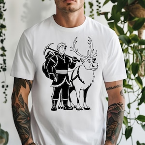 Op de afbeelding: Wit t-shirt met een zwart-witte afbeelding van een personage met een pikhouweel en een rendier. Het personage draagt een donkere outfit en draagt een tas. Het rendier heeft een harnas.
