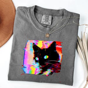 Può includere: Maglietta grigia con un disegno di gatto nero in stile glitch art colorato. Gli occhi del gatto sono verdi e blu. Una collana d'argento con un piccolo ciondolo è appoggiata sulla maglietta. La maglietta è piegata su una superficie bianca.