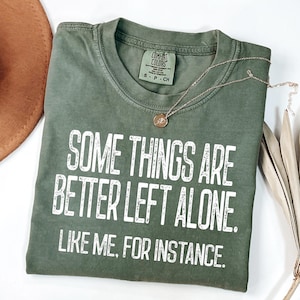 May include: Olive green t-shirt with the text "SOME THINGS ARE BETTER LEFT ALONE. LIKE ME, FOR INSTANCE." A gold necklace with a pendant is draped across the shirt. The shirt is folded on a white surface.
