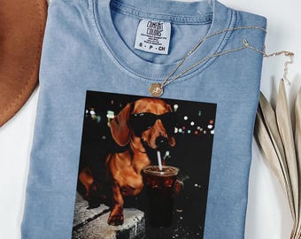 Può includere: Una maglietta Comfort Colors azzurra con una grafica di un bassotto con occhiali da sole che beve da una tazza con una cannuccia. Una collana d'oro è drappeggiata sulla maglietta. La maglietta è una taglia S-P-CH.