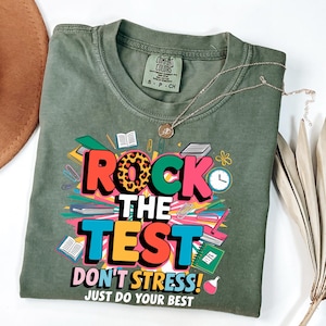 Op de afbeelding: Olijfgroen t-shirt met de tekst "ROCK THE TEST" in kleurrijke blokletters. Het ontwerp bevat boeken, potloden en een klok. Het shirt heeft ook de zinnen "DON'T STRESS!" en "JUST DO YOUR BEST".