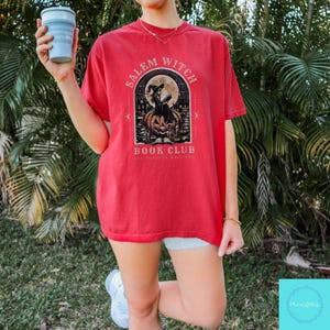 May include: Red t-shirt with a graphic featuring a witch reading a book in front of a full moon, with the text "Salem Witch Book Club". The design includes a jack-o'-lantern and other Halloween-themed elements. The shirt is a short-sleeved style.