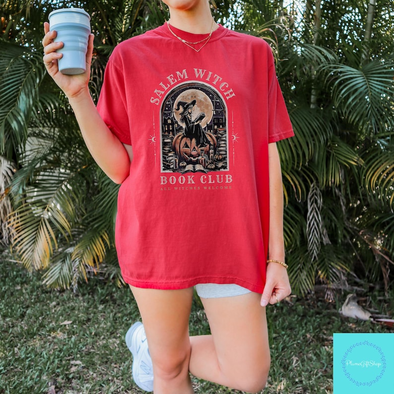 May include: Red t-shirt with a graphic featuring a witch reading a book in front of a full moon, with the text "Salem Witch Book Club". The design includes a jack-o'-lantern and other Halloween-themed elements. The shirt is a short-sleeved style.