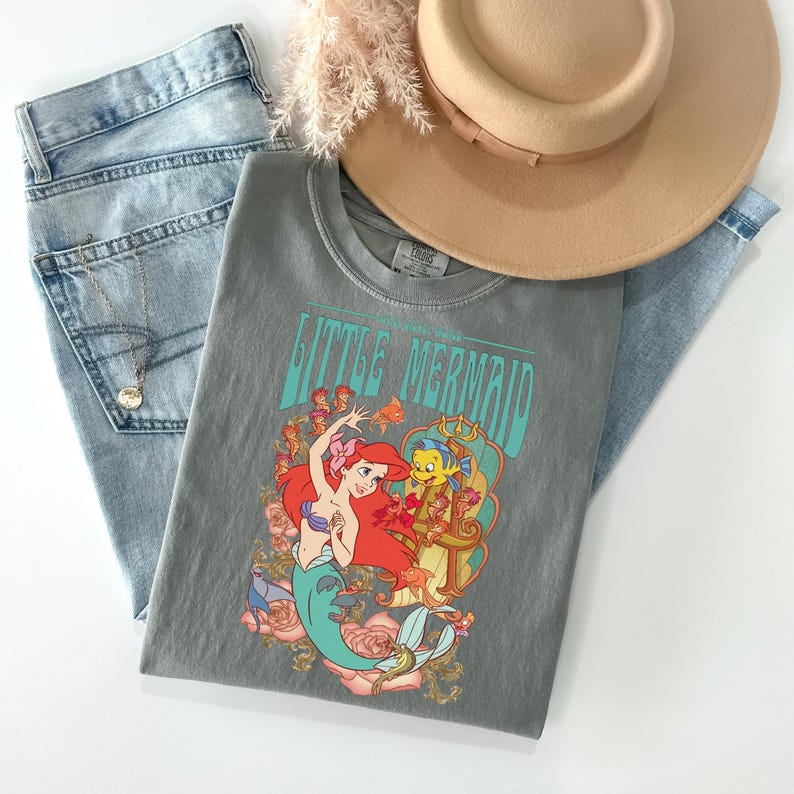 May include: A gray t-shirt featuring the text "Little Mermaid" with a graphic of Ariel and Flounder. The shirt is paired with light wash denim jeans, a tan hat, and a gold necklace. A sprig of dried flowers is also included.