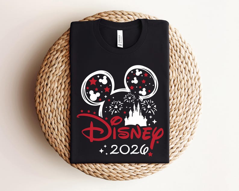 May include: Black t-shirt featuring a white and red Disney design. The design includes Mickey Mouse ears, stars, fireworks, a castle, and the word "Disney" in red, along with the year "2026". The shirt is displayed on a woven surface.