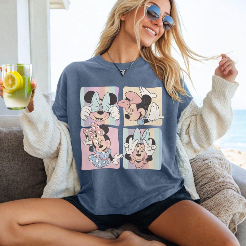 Op de afbeelding: Blauw T-shirt met vier panelen met verschillende Minnie Mouse-illustraties. Elk paneel heeft een pastelkleurige regenboogachtergrond. Minnie draagt verschillende strikken en poses. Het T-shirt heeft een relaxte pasvorm.