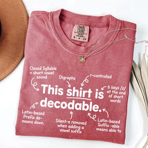 May include: A dusty rose short-sleeved t-shirt featuring white text that reads This shirt is decodable, surrounded by linguistic notes including Closed Syllable, Digraphs, r-controlled, Latin-based Prefix de-, Silent-e, and Latin-based Suffix -able.