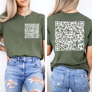 May include: An olive green t-shirt featuring a white QR code graphic on the front left chest and a larger white QR code graphic printed on the back. The shirt has short sleeves and a crew neckline, paired with blue denim jeans with distressed details.