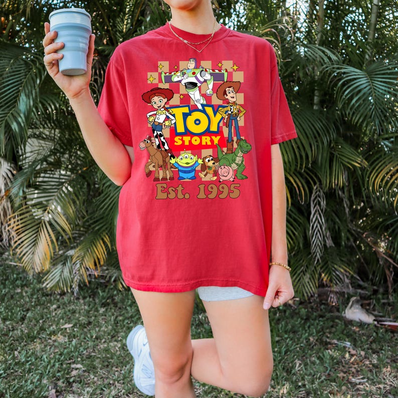 May include: Red t-shirt featuring the Toy Story logo and characters, including Woody, Buzz Lightyear, and Jessie. The shirt also has the text "Est. 1995". The shirt is a comfortable, casual style.