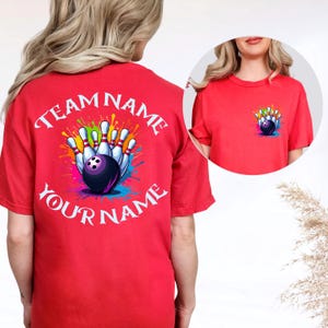 Może przedstawiać: Czerwona koszulka z grafiką o tematyce kręglarskiej. Projekt przedstawia kulę do kręgli i kręgle z kolorowymi rozpryskami farby oraz tekst "TEAM NAME" i "YOUR NAME". Mniejsza wersja grafiki znajduje się z przodu.