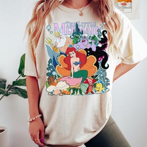 May include: Cream-colored t-shirt featuring a colorful graphic of characters from "The Little Mermaid." The design includes Ariel, King Triton, Ursula, and Flounder. The words "The Little Mermaid" are at the top in a stylized font.