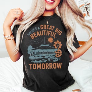 May include: Black t-shirt featuring a retro-style graphic with the text "There's a Great Big Beautiful Tomorrow." The design includes a futuristic building, a sun, and gears in shades of orange and blue. A vintage-inspired tee.