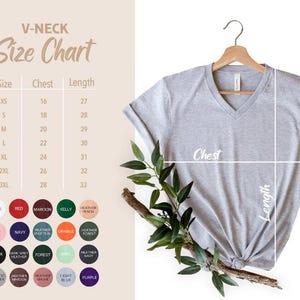 Op de afbeelding: Een lichtgrijs V-hals T-shirt hangt aan een houten hanger, met een maattabel en kleuropties aan de linkerkant. De tabel toont borst- en lengtematen in cm. Groene bladeren en een tak zijn voor het shirt geplaatst.