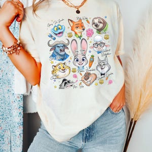 May include: A cream-colored short-sleeve t-shirt featuring a colorful graphic print of various animated animal characters from Zootopia, including Judy Hopps, Nick Wilde, and Flash, along with small icons like carrots, paw prints, and police badges.