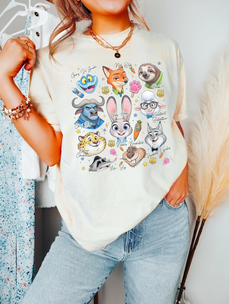 May include: A cream-colored short-sleeve t-shirt featuring a colorful graphic print of various animated animal characters from Zootopia, including Judy Hopps, Nick Wilde, and Flash, along with small icons like carrots, paw prints, and police badges.