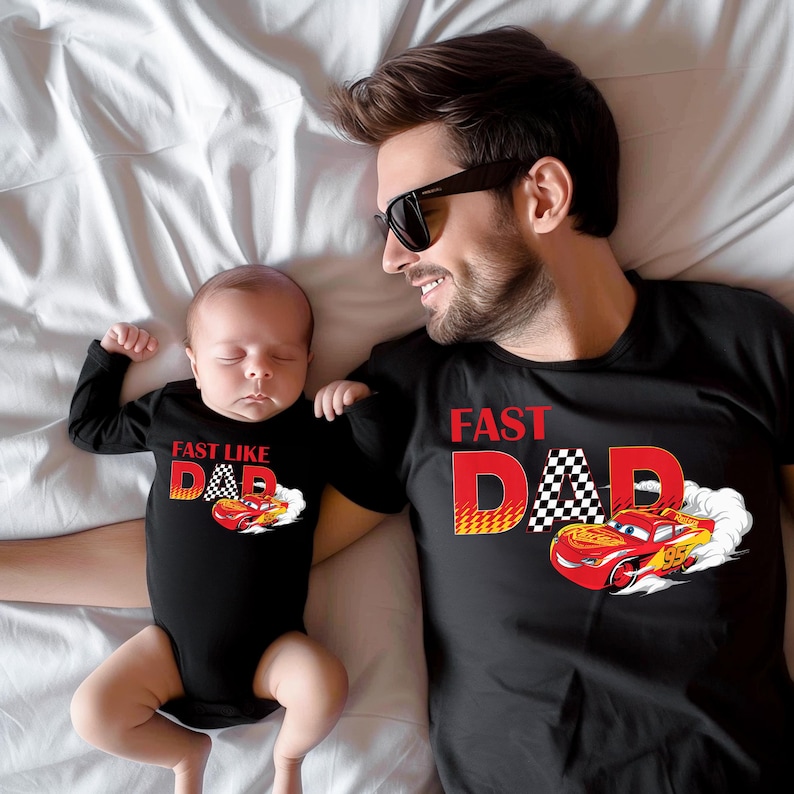 May include: Black t-shirts and a baby bodysuit with a race car theme. The adult's shirt says "Fast Dad" with a checkered flag and a red race car. The baby's bodysuit says "Fast Like Dad" with a similar design.