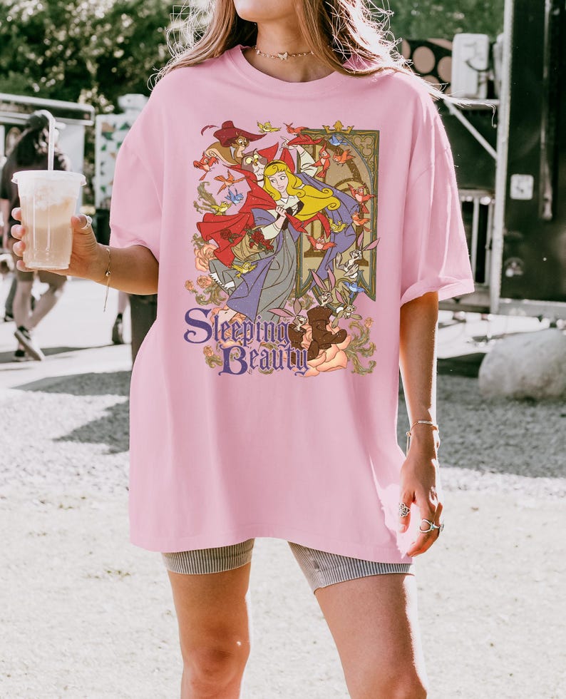 May include: A light pink short-sleeve t-shirt featuring a graphic print of the character Aurora from Sleeping Beauty, surrounded by forest animals and birds. The text Sleeping Beauty is printed in a purple serif font at the bottom of the design.