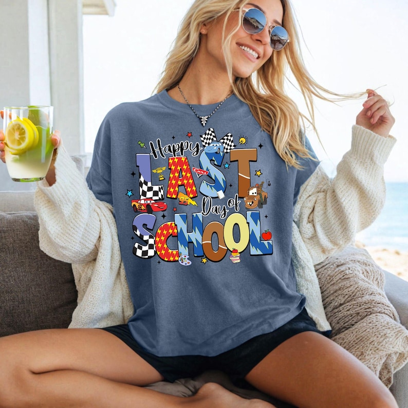 Peut inclure: Un t-shirt bleu avec l'inscription "Happy Last Day of School" dans des lettres color&eacute;es et ludiques. Le motif comprend des voitures de course, un drapeau &agrave; damier et une voiture de dessin anim&eacute;. Le t-shirt est port&eacute; par une personne portant des lunettes de soleil.