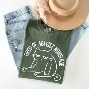 May include: An olive green t-shirt featuring a white graphic of a grumpy cat and the text "TIRED OF ABLEIST NONSENSE" in a curved font. The shirt is layered with light blue denim jeans and a tan wide-brimmed hat with a ribbon band.