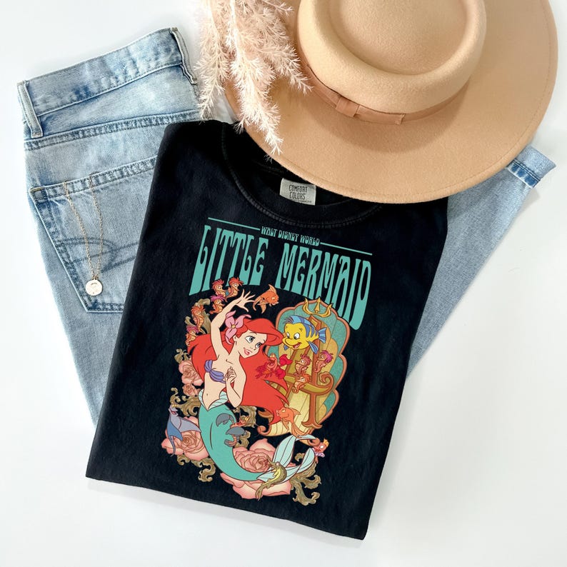 May include: Black t-shirt featuring a colorful graphic of Ariel and Flounder from The Little Mermaid, with the text "Little Mermaid." The shirt is paired with light-wash jeans and a tan hat. A gold necklace and dried flowers complete the casual ensemble.