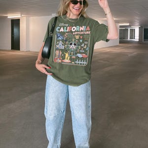 Op de afbeelding: Olijfgroen T-shirt met een kleurrijke afbeelding van Disney California Adventure. Gedragen met lichtblauwe jeans en witte sneakers. Een zwarte schoudertas maakt de outfit compleet.