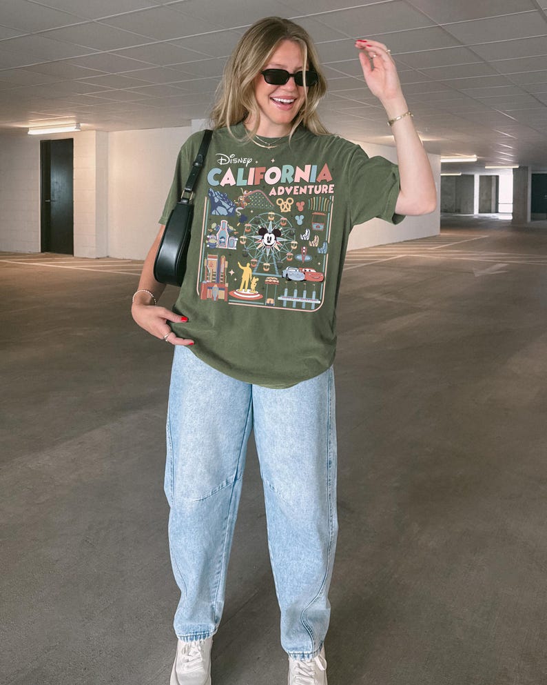 Op de afbeelding: Olijfgroen T-shirt met een kleurrijke afbeelding van Disney California Adventure. Gedragen met lichtblauwe jeans en witte sneakers. Een zwarte schoudertas maakt de outfit compleet.