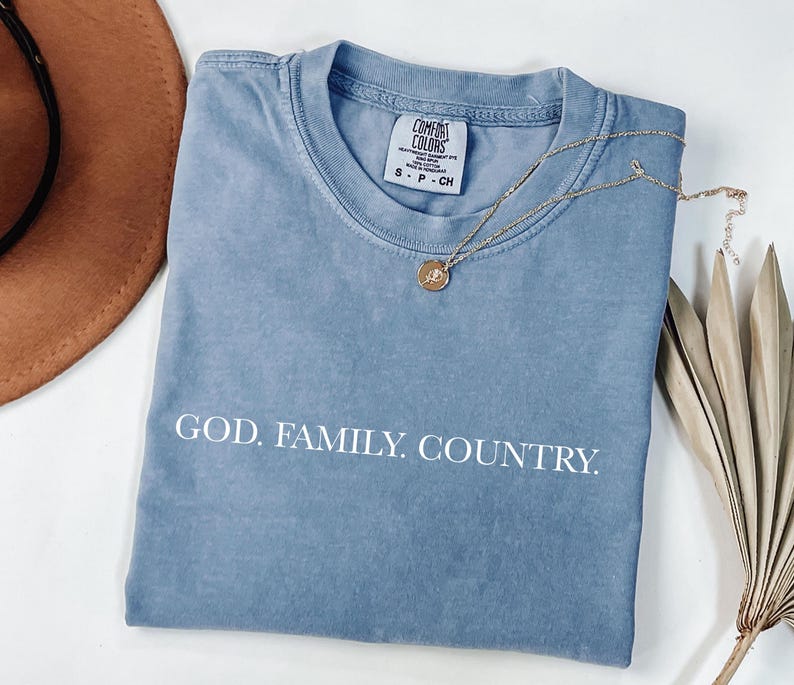 May include: A blue Comfort Colors t-shirt with the words "GOD. FAMILY. COUNTRY." printed in white. A gold necklace with a pendant rests on the shirt. A brown hat and dried plant are in the background.