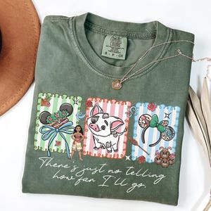 May include: An olive green t-shirt featuring a graphic print of three rectangular panels with Moana, Pua the pig, and character-themed mouse ears. The text "There's just no telling how far I'll go" is printed in white script below the illustrations.