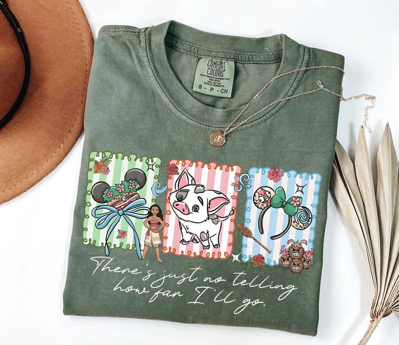 May include: An olive green t-shirt featuring a graphic print of three rectangular panels with Moana, Pua the pig, and character-themed mouse ears. The text "There's just no telling how far I'll go" is printed in white script below the illustrations.