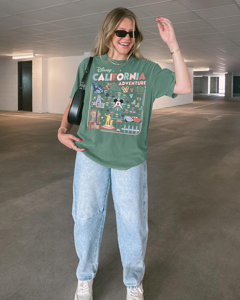 Op de afbeelding: Groen t-shirt met een afbeelding van Disney California Adventure. Het shirt heeft Mickey Mouse en verschillende parkattracties. De persoon draagt een lichtblauwe spijkerbroek en witte sneakers. De persoon draagt ook een zwarte zonnebril en een zwarte handtas.
