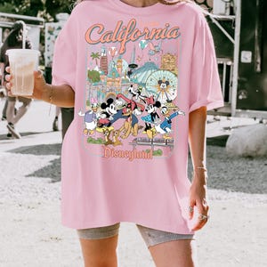 Op de afbeelding: Roze T-shirt met een Disneyland-afbeelding. De afbeelding toont Mickey Mouse, Minnie Mouse, Donald Duck en Goofy voor Disneyland-bezienswaardigheden. De tekst "California Disneyland" staat bovenaan de afbeelding.
