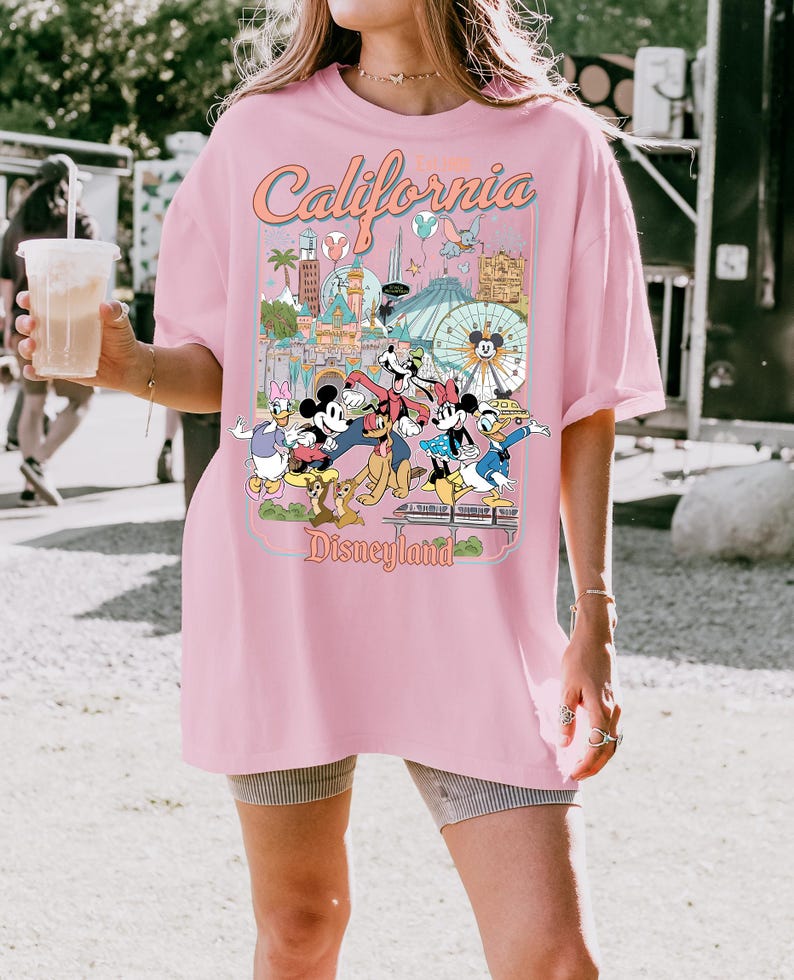 Op de afbeelding: Roze T-shirt met een Disneyland-afbeelding. De afbeelding toont Mickey Mouse, Minnie Mouse, Donald Duck en Goofy voor Disneyland-bezienswaardigheden. De tekst "California Disneyland" staat bovenaan de afbeelding.