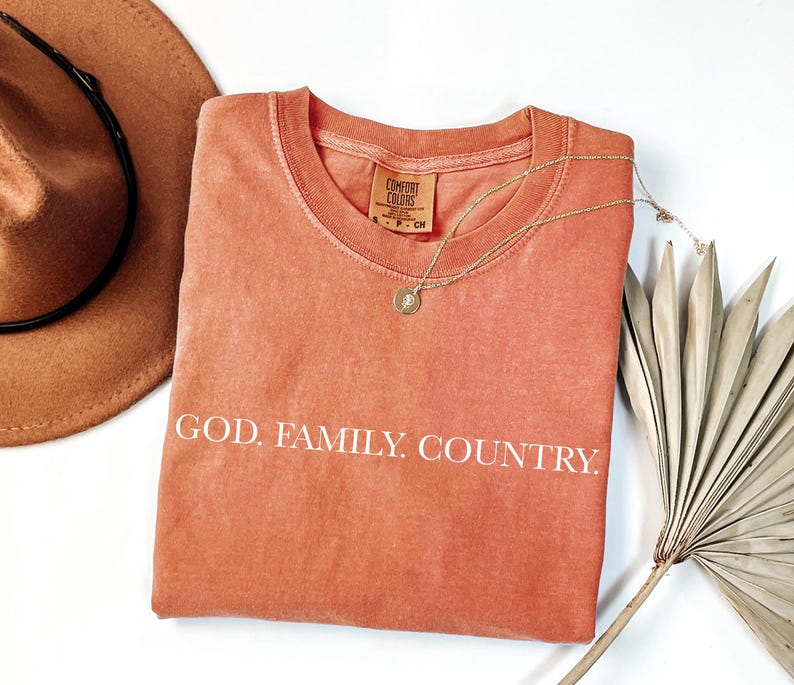 May include: A coral-colored Comfort Colors t-shirt with the words "GOD. FAMILY. COUNTRY." in white. A gold necklace with a pendant and a brown hat are also in the image. The shirt has a crew neck and short sleeves.
