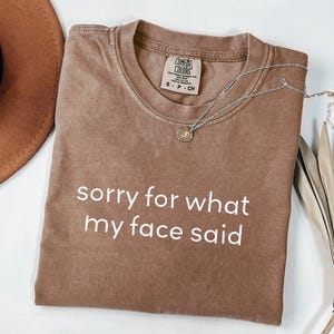 May include: A brown Comfort Colors t-shirt with the text "sorry for what my face said" in white. A silver necklace with a small pendant is draped across the shirt. A brown hat and dried palm leaves are in the background.