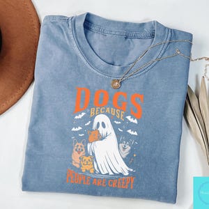 Może przedstawiać: Niebieska koszulka z pomarańczowym napisem "DOGS BECAUSE PEOPLE ARE CREEPY". Grafika przedstawia ducha trzymającego psa, z innymi psami i nietoperzami. Złoty naszyjnik jest zarzucony na koszulkę.