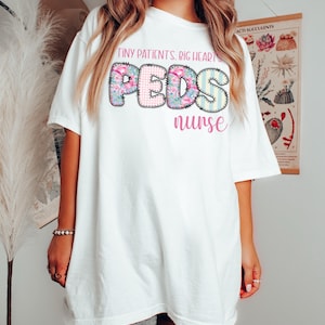 May include: White t-shirt with the text "TINY PATIENTS. BIG HEARTS. PEDS nurse." The word "PEDS" is in a colorful, floral and striped design. The shirt is a casual, oversized fit.