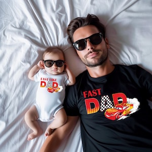 May include: A father and baby wearing sunglasses and matching apparel. The baby wears a white onesie with the text "FAST LIKE DAD" and a race car graphic. The father wears a black t-shirt with the same design.
