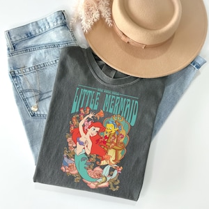 May include: A gray t-shirt featuring a colorful graphic of Ariel and Flounder from The Little Mermaid, with the text "LITTLE MERMAID". The shirt is laid on top of light blue jeans, a tan hat, and a gold necklace.