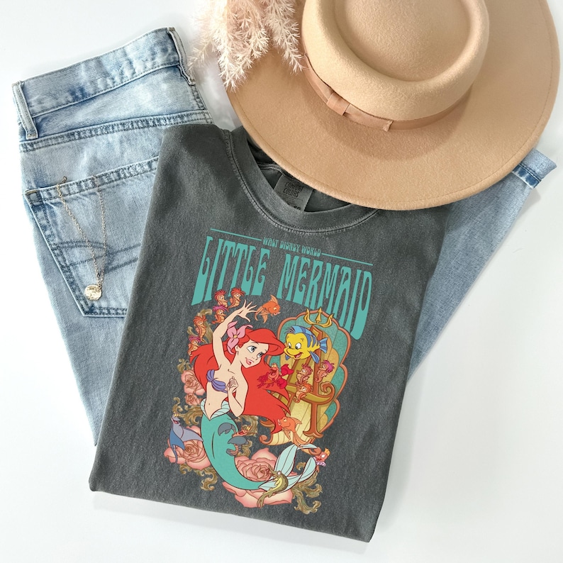 May include: A gray t-shirt featuring a colorful graphic of Ariel and Flounder from The Little Mermaid, with the text "LITTLE MERMAID". The shirt is laid on top of light blue jeans, a tan hat, and a gold necklace.
