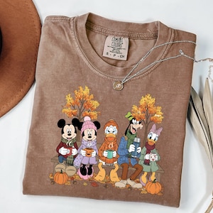May include: A brown short-sleeved t-shirt featuring a graphic print of Mickey Mouse, Minnie Mouse, Donald Duck, Goofy, and Daisy Duck sitting on a bench in an autumn setting. The characters hold mugs and a thermos. The shirt has a Comfort Colors label.